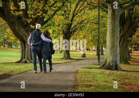 Couple walking together on scenic park path (arms round each other) by  trees (colourful autumn foliage or leaves) - The Stray, Harrogate England UK Stock Photo