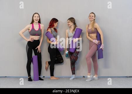 four charming athletic girls in sports out fits talking and smiling standing next to the wall holding a yoga mat, smiling to each other, standing at a Stock Photo