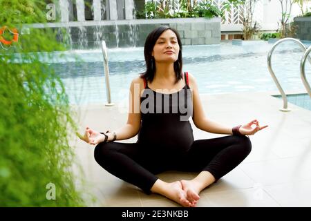 Pregnant Southeast Asian Woman in Black Tank Top and Legging Sitting Cross-Legged By Poolside Doing Yoga Exercise for Health Stock Photo