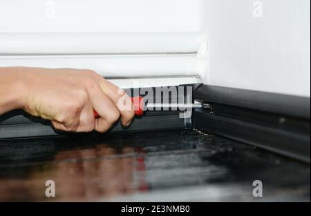 A man is installing black plastic skirting board to a white wallpapered wall in the corner of a room with central heating pipes using a screwdriver. Stock Photo