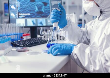 Medical researcher developing cure for coronavirus working with micropipette and petri dish dressed in coverall. Chemist in modern laboratory doing research using dispenser during global epidemic with covid-19. Stock Photo