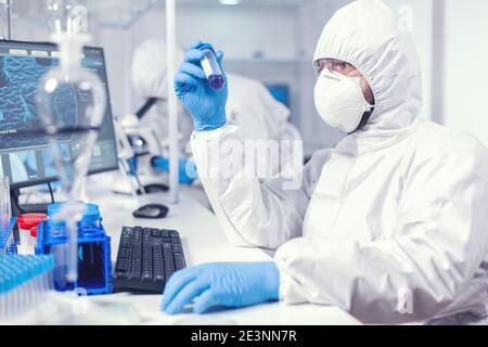 Lab technician dressed in protective suit as safety precaution looking at test tube. Scientist in laboratory wearing coverall doing research and analyzing substance during global pandemic with covid19. Stock Photo