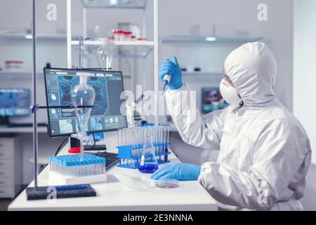 Researcher analyzing sample in automatic pipette working in lab dressed in coverall. Chemist in modern laboratory doing research using dispenser during global epidemic with covid-19. Stock Photo