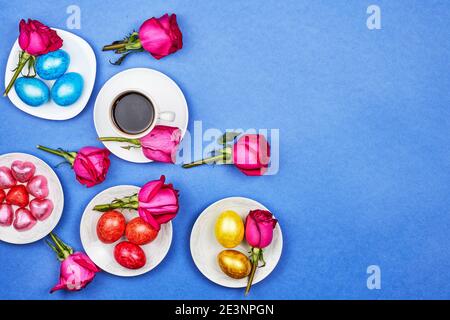Easter eggs, red rose flowers, a cup of black coffee on blue background Stock Photo