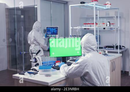 Scientist conducting experiment using computer with green screen sitting his workpalce in lab. Team of microbiologists doing vaccine research writing on device with chroma key, isolated, mockup display. Stock Photo