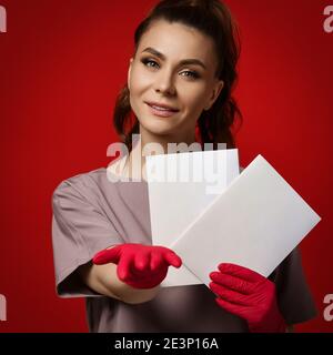 Young smiling woman doctor therapist in protective latex gloves holding two paper sheets blanks, pointing and looking at camera Stock Photo