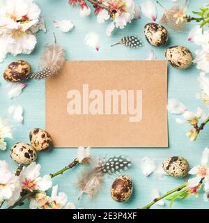 Easter holiday background. Flat-lay of Spring almond blossom flowers on branches, feathers, quail eggs, paper in center over blue background, top view, copy space, square crop. Greeting card concept Stock Photo