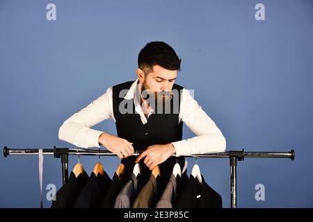 Man with beard in vest by clothes rack. th curious face near custom jackets on blue background. Business fashion and sewing concept. Designer leans on clothes hangers with suits. Stock Photo