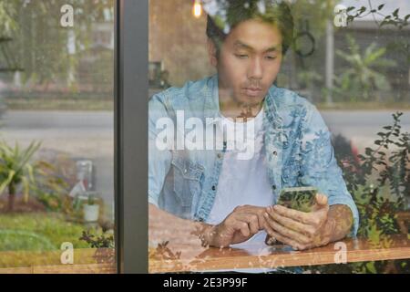 Young Asian Casual Businessman in Denim or Jeans Shirt Using Smartphone in Coffee Shop. Casual Businessman Working with Technology Stock Photo