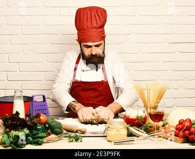 Cook with concentrated face in burgundy uniform sits by table with vegetables and kitchenware. Cooking process concept. Chef makes dough. Man with beard kneads dough on white brick wall background. Stock Photo