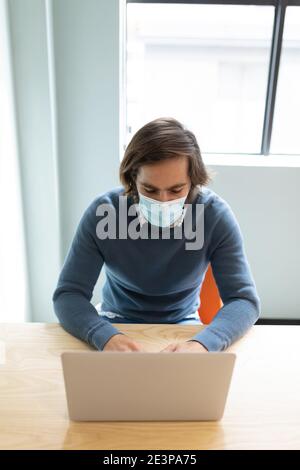 Caucasian businessman wearing face mask sitting at desk using laptop Stock Photo