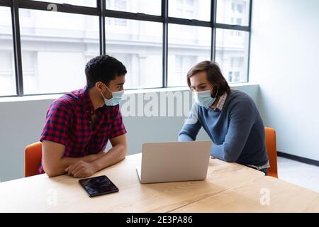 Two diverse businessmen wearing face masks using laptop and discussing in creative office Stock Photo