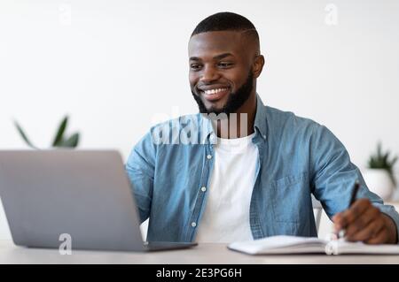 Black Millennial Male Entrepreneur Taking Notes While Working On Laptop In Office Stock Photo
