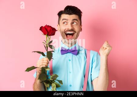 Romance and Valentines day concept. Cheerful man in bow-tie screaming from happiness, holding red rose and jumping on date, standing over pink Stock Photo