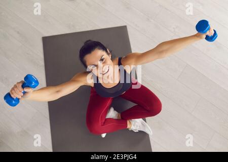 Sporty woman sitting on gym mat, exercising with dumbbells and smiling at camera Stock Photo