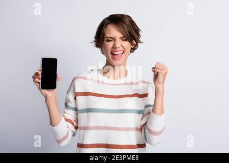 Photo of lucky cute young lady wear striped sweater holding modern gadget rising fist closed eyes isolated grey color background Stock Photo