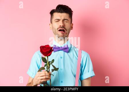 Heartbroken guy in funny bow-tie crying over girlfriend, standing alone with red rose on pink background and sobbing, break-up on Valentines day Stock Photo
