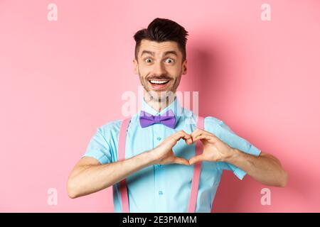Passionate guy in funny bow tie saying I love you, showing heart gesture on Valentines day and smiling, expressing sympathy to lover, standing over Stock Photo