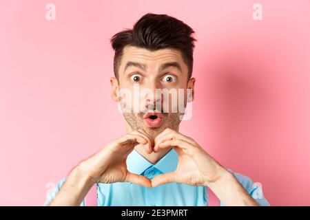 Valentines day concept. Close up of handsome caucasian man falling in love, looking amazed and showing heart gesture, pink background Stock Photo