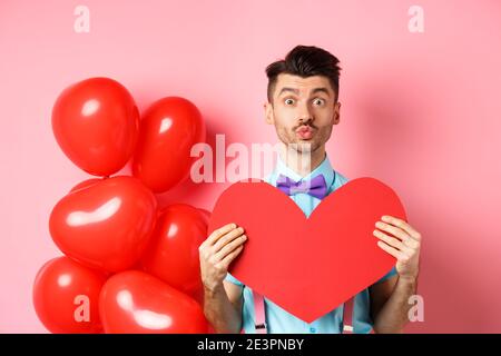 Valentines day concept. Cute guy pucker lips for kiss and showing romantic heart cutout, falling in love, standing over pink background Stock Photo