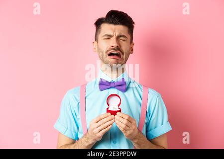 Valentines day. Heartbroken guy crying and holding engagement ring, sobbing from break-up, standing over pink background Stock Photo