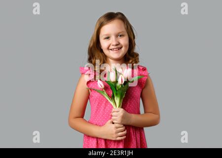 Cute happy little girl in a pink dress with polka dots on a gray background. The child is holding a bouquet of tulips. Stock Photo