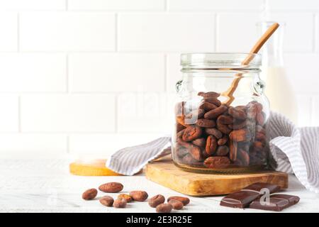 Cocoa beans in a glass jar on a light kitchen table. Culinary food cooking background for making traditional chocolate. Selective focus. Stock Photo
