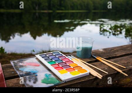 Close-up of watercolor setup with aquarelle palette paintbrushes and water jar lay on a wooden bench by a forest lake - plein air scetching concept Stock Photo