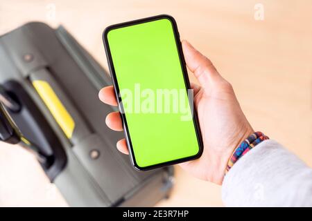 (Selective focus) Overhead view of an human hand holding a smart phone with a green screen with a defocused luggage in the background. Stock Photo