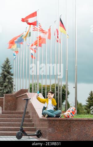 Teenager sits near the administration building against background of country flags and waves into the smartphone. Electric scooter rider concept Stock Photo