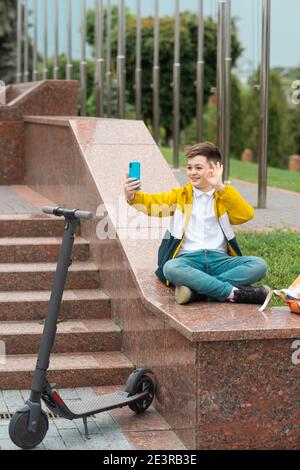 Teenager sits near the administration building against background of country flags and waves into the smartphone. Electric scooter rider concept Stock Photo