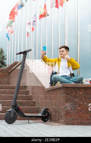 Teenager sits near the administration building against background of country flags and waves into the smartphone. Electric scooter rider concept Stock Photo