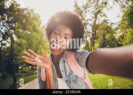 Photo portrait of curly black skinned beautiful girl taking selfie in park smiling waving with palm wearing casual jeans shirt backpack
