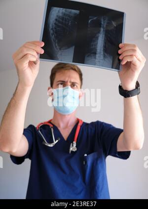 a handsome physician men wearing scrub, stethoscope and face mask looking to an x-ray during the sars cov 2 pandemic covid 19 Stock Photo