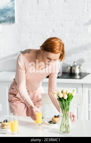 redhead tattooed woman serving breakfast on table near tulips in kitchen Stock Photo