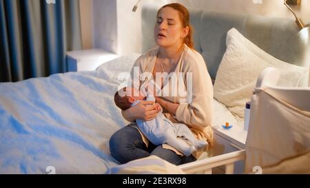 Tired mother rocking and feeding her newborn baby on bed at night. Concept of healthy and natural baby nutrition. Health of mother and child. Stock Photo
