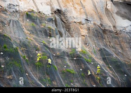 Workers strengthen the slope of the mountain with a metal mesh preventing rockfall on the road. Hondarribia, Spain. Abstract safety concept background Stock Photo