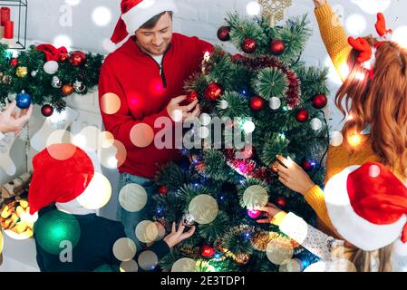 happy family in santa hats decorating christmas tree together at home, bokeh lights illustration Stock Photo