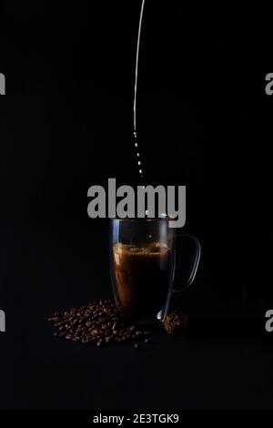 Milk is pouring into black coffee in glass cup on old dark background with beans and chocolate truffle. Breakfast coffee love concept. Front view Stock Photo