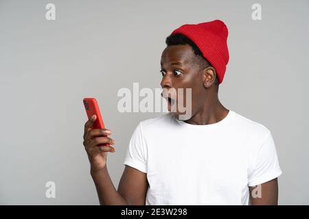 Surprised afro man makes big eyes opens mouth widely, shocked with indecent photo, looking at mobile Stock Photo