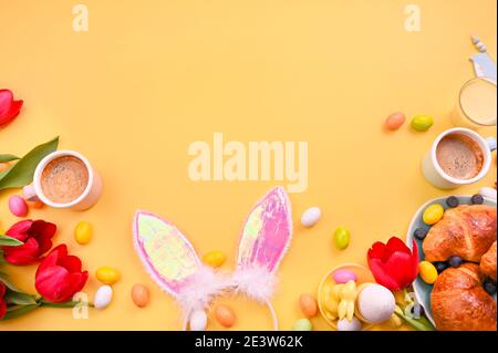 Spring breakfast. Espresso coffee in bright cups, Easter chocolate eggs, pink bunny ears and red tulips on a yellow background. Free space for text. Copy space. Flat lay  Stock Photo