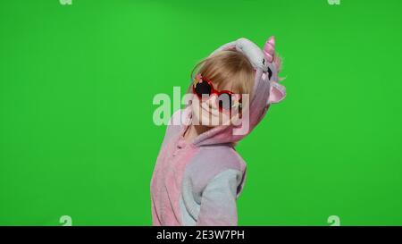 Portrait of a cute young little child in sunglasses smiling, dancing in unicorn costume on chroma key green background. Kid girl animator making gun gesture with hands in unicorn pajamas. Copy space Stock Photo