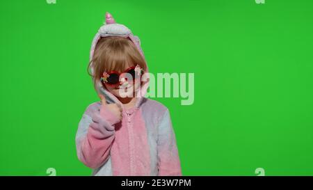 Portrait of a cute young little child in sunglasses smiling, dancing in unicorn costume on chroma key green background. Kid girl animator showing thumbs up with hands in unicorn pajamas. Copy space Stock Photo