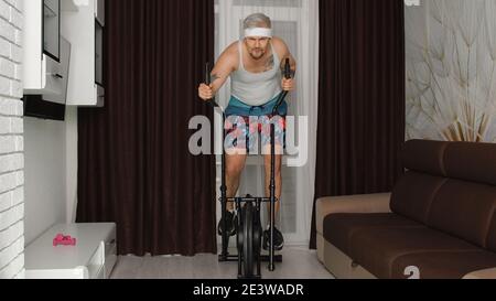 Funny Caucasian athlete sportsman guy couch on orbitrek in living room. Stylish hipster man blogger trainer recording sport fitness cardio online vlog at home. Sportive humor. Joke, comical, parody Stock Photo