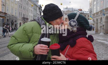 Senior couple tourists grandmother grandfather making a kiss, drinking from plastic cups, enjoying hot drink mulled wine, tea, coffee in city center. Elderly pensioners family Christmas vacations Stock Photo