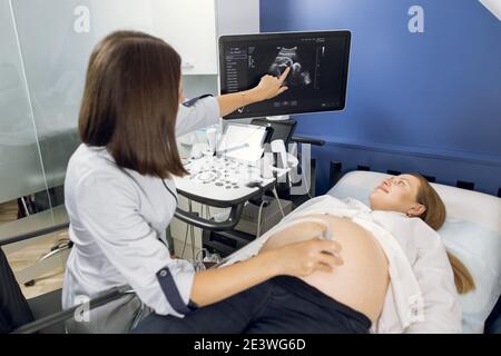 Back view of female ultrasound technician, scanning pregnant woman's belly in modern clinic, pointing on the screen with baby's image Stock Photo