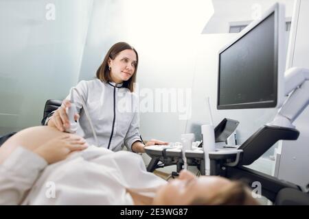 Young smiling professional attractive woman doctor doing 3d ultrasound on belly of pregnant woman in clinic. Ultrasound for pregnant woman Stock Photo