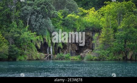 Closeup on waterfall and cave on the edge of turquoise lake. Green lush forest in Plitvice Lakes National Park UNESCO World Heritage, Croatia Stock Photo