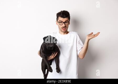 Image of handsome young man holding black pug and looking confused. Guy shrugging shoulders and staring indecisive at camera, carry dog in arm, white Stock Photo