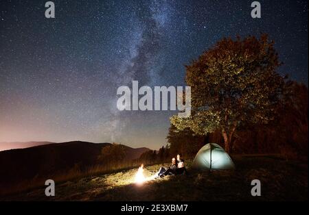 Romantic couple travellers having a rest at campfire beside camp and tourist tent under amazing night sky full of stars and Milky way. On background beautiful starry sky, mountains and big tree Stock Photo